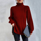 Woman wearing an oversized red turtleneck knit sweater with long sleeves, textured waffle pattern, and side slits, styled with a black quilted handbag.