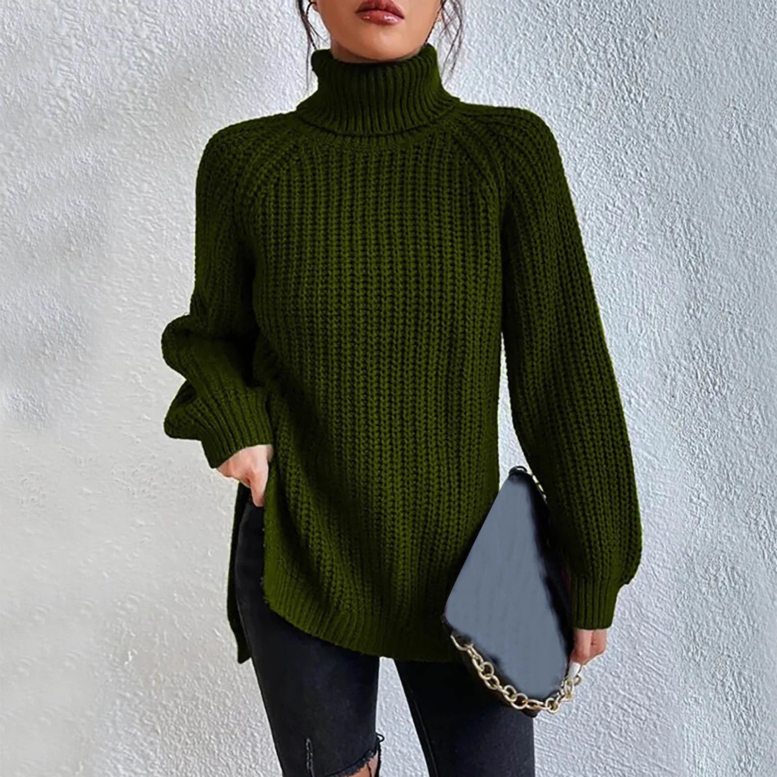 Woman wearing an oversized dark green turtleneck knit sweater with long sleeves, textured waffle pattern, and side slits, styled with a black quilted handbag.