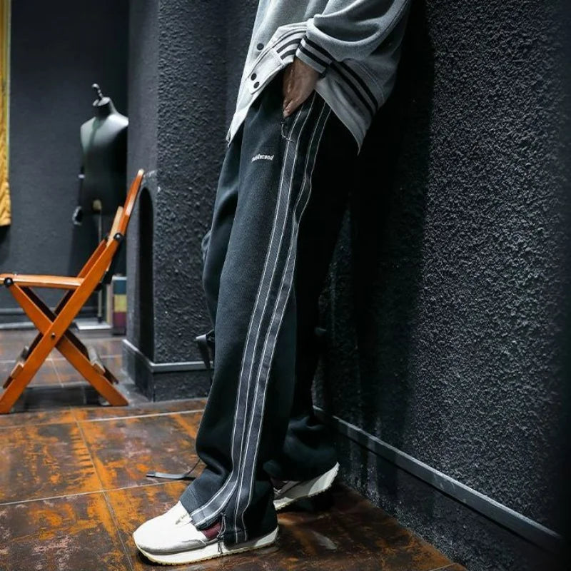 Man wearing black track pants with white side stripes, leaning against wall.