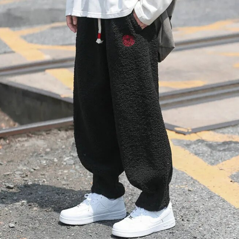 Person wearing loose black fleece jogger pants with drawstring waist and red embroidery detail, paired with white sneakers on a street.