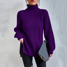 Woman wearing an oversized purple turtleneck knit sweater with long sleeves, textured waffle pattern, and side slits, styled with a black quilted handbag.