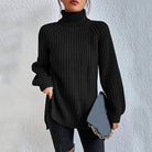 Woman wearing an oversized black turtleneck knit sweater with long sleeves, textured waffle pattern, and side slits, styled with a black quilted handbag.