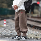 Person wearing loose brown fleece jogger pants with drawstring waist and red embroidery detail, paired with white sneakers on a street.