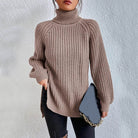 Woman wearing an oversized khaki turtleneck knit sweater with long sleeves, textured waffle pattern, and side slits, styled with a black quilted handbag.